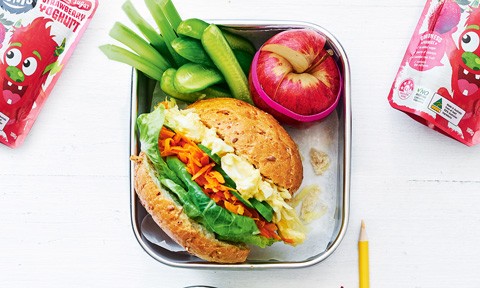 An egg and salad roll in a lunch box with an apple and celery