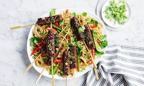  Korean Beef Skewers on Noodles, placed on serving plate on marble table. Spring onion on the dish and on side plate, arranged next to a striped towel.