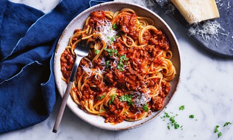 Curtis Stone's Spaghetti Bolognese served in a ceramic bowl, topped with parsley and parmesan. A blue table cloth is on a marble background, a block of Parmesan on a navy plate.