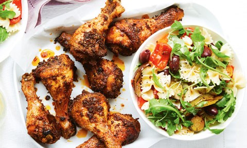 Chicken drumsticks served on a baking tray alongside a bowl of pasta salad with greens, tomatoes and olives