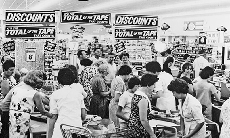 Black and white image of crowded checkout and bagging area and large discount banners