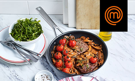 Frying pan with pasta and tomatoes on a kitchen table with cutlery and fresh herbs beside