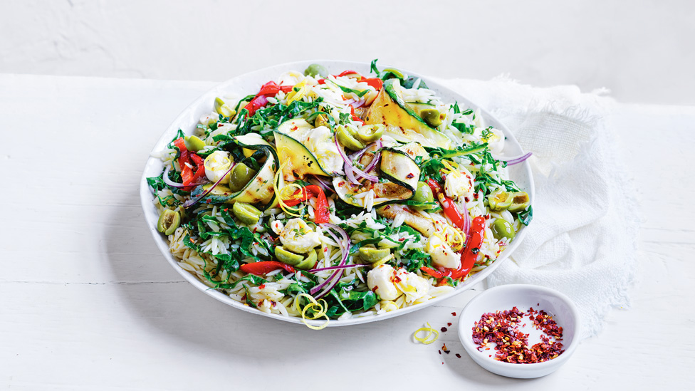 Antipasto salad with bocconcini, risoni, courgette ribbons and red capsicums served in a bowl with chilli flakes beside it