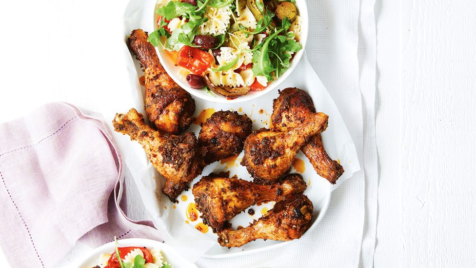 Chicken drumsticks served on a baking tray alongside a bowl of pasta salad with greens, tomatoes and olives