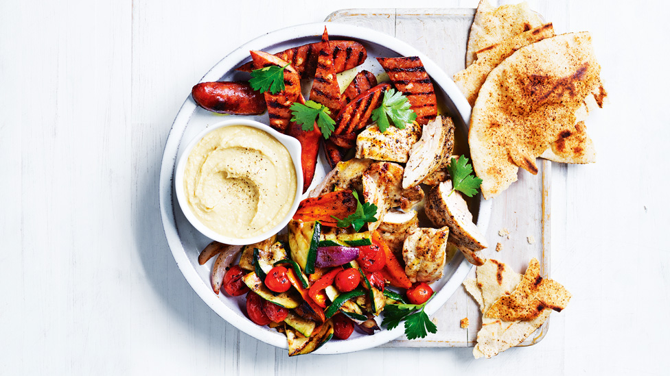 Large plate with chopped chicken, chorizo, chargrilled veggies, topped with herbs and a small bowl of hommus, with torn flatbread on side