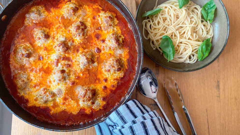 A pan of cheesy, oven-baked meatballs with pasta and basil leaves.