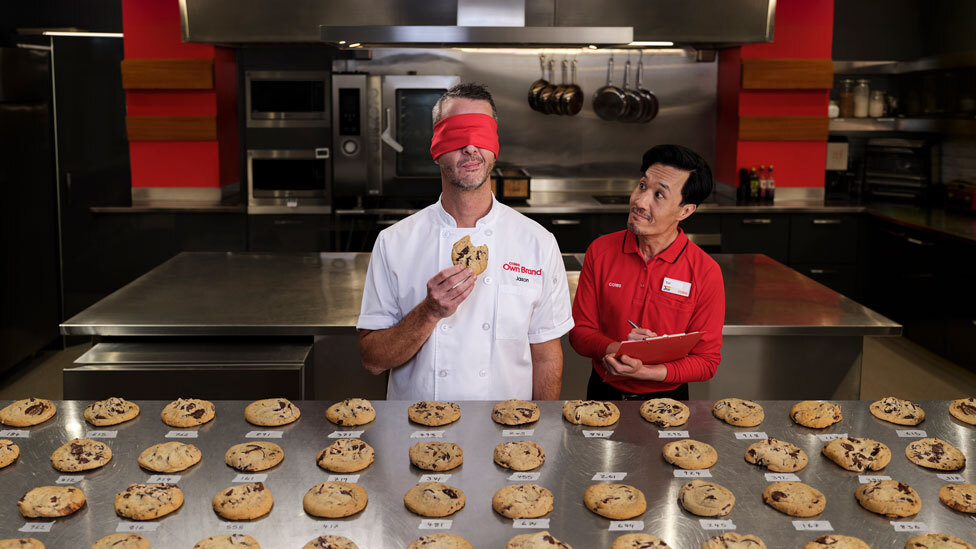 Man tasting cookies blindfolded while another man smiles and takes notes