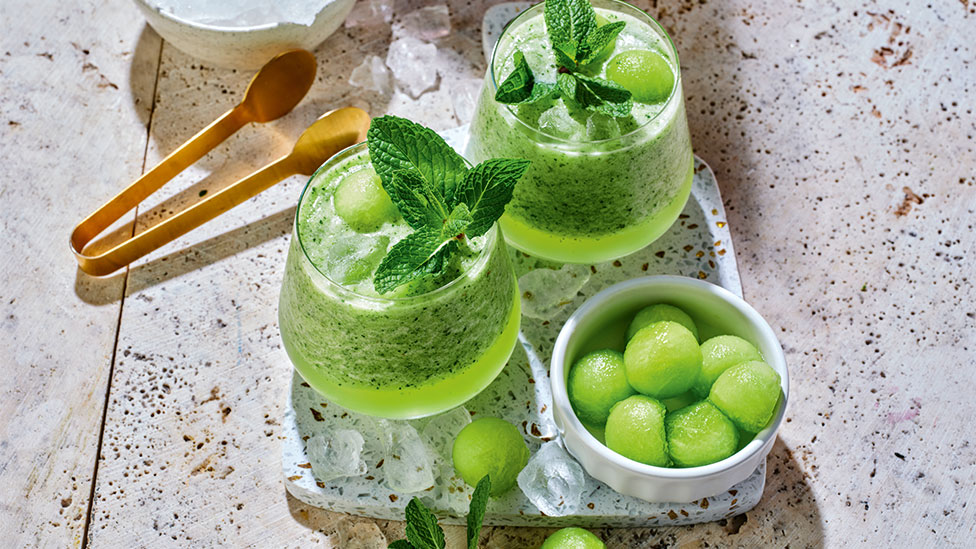 Two green melon mojitos with green meon balls in an adjacent bowl