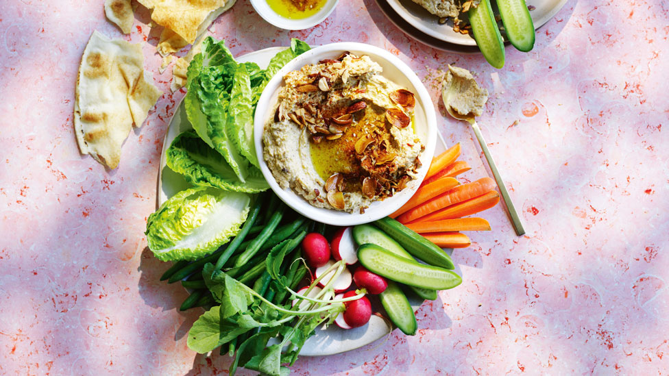 Roast cauliflower hommus in a bowl with vegetable sticks around it on a plate