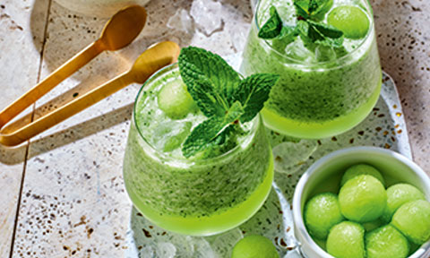 Two green melon mojitos with green meon balls in an adjacent bowl