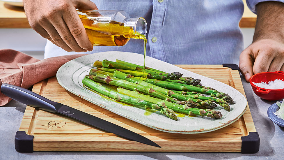 Asparagus on a white serving platter, being drizzled with oil.