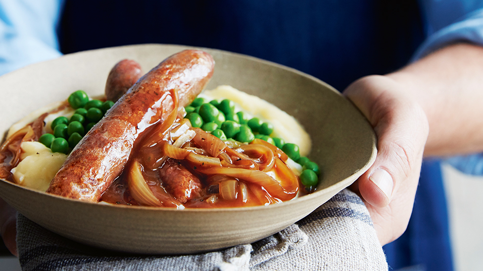Sausages, onions in gravy, peas and mashed potato in a bowl