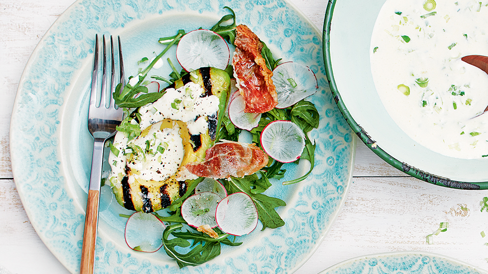 char grilled avocado half on a plate with salad greens and crispy prosciutto