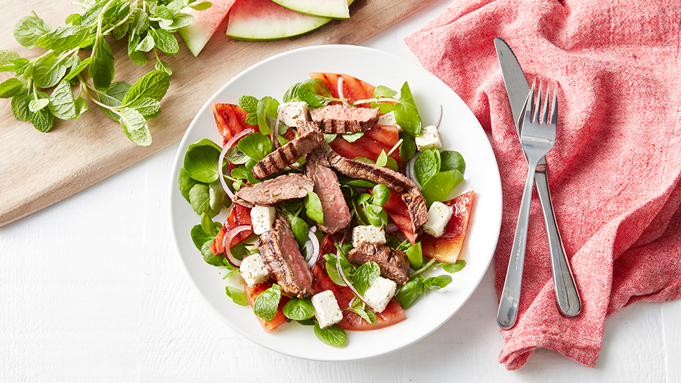 A salad of sliced steak, watermelon, feta and greens in a bowl