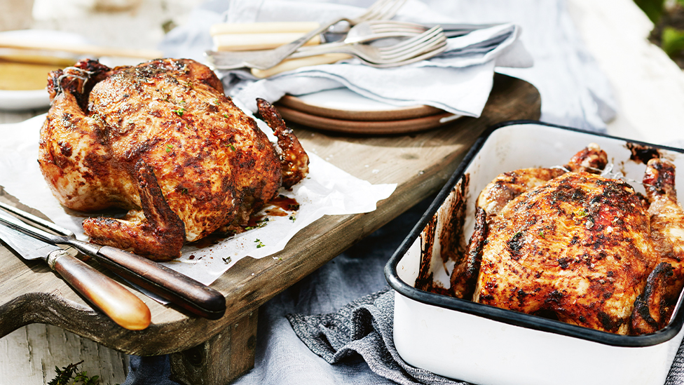 Two whole barbecued chickens, one on a board and the other in a baking dish