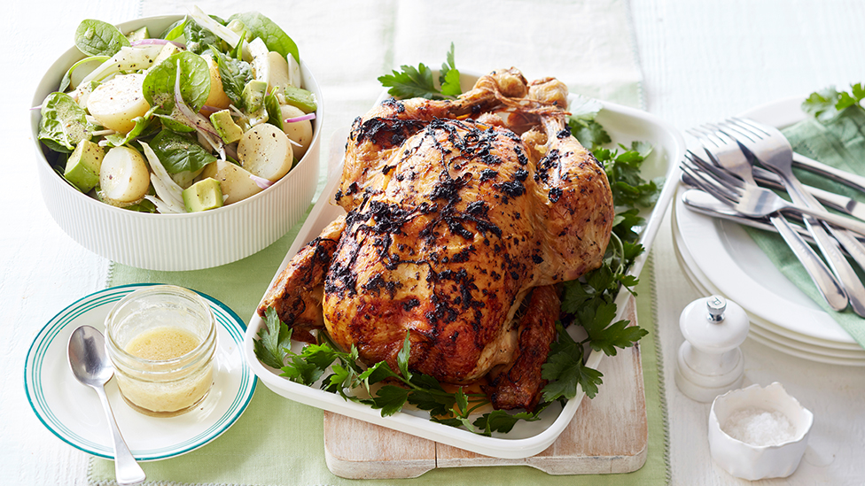 Whole barbecued chicken in a dish alongside a potato and baby spinach salad in bowl