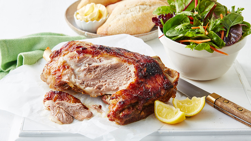 Roasted lamb shoulder on a board accompanied by a salad of greens in a bowl and bread rolls