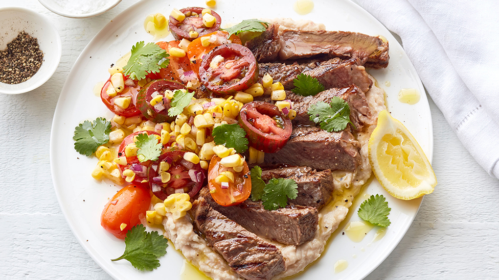 Sliced barbecued steak served on white bean puree, with a tomato and corn salad