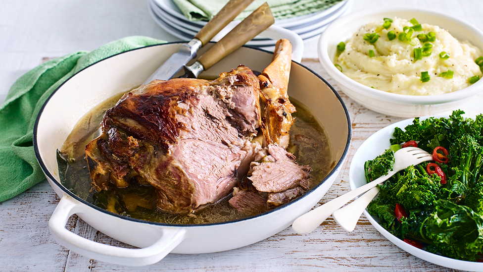 Roast leg of lamb in a baking dish, with mashed potato and greens in dishes on the side