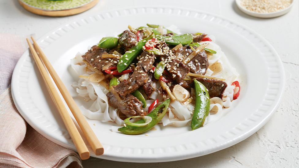 Plate of sliced steak with sugar snap peas and red chilli on wide rice stick noodles, with chop sticks to the side