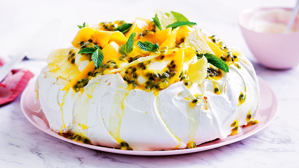 Pavlova on a plate topped with cream, mango, pineapple, passionfruit pulp and mint leaves