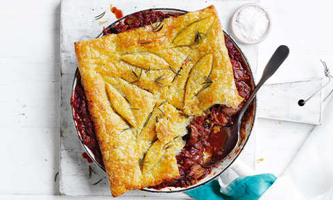 Slow-cooked Lamb Shank Pie in oven dish topped with oregano,. Placed on a white wooden board with a side of coarse salt.