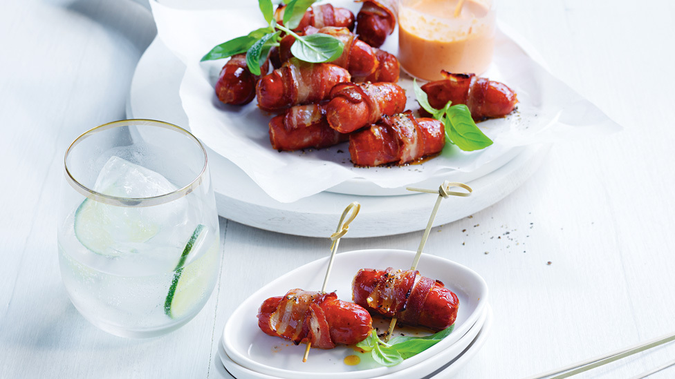 A batch of bacon-wrapped chorizo garnished with basil leaves, with capsicum mayo on the side.