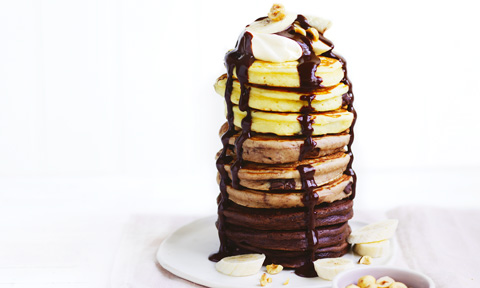 A stack of tri coloured pancakes topped with chocolate sauce, banana, cream and hazelnuts