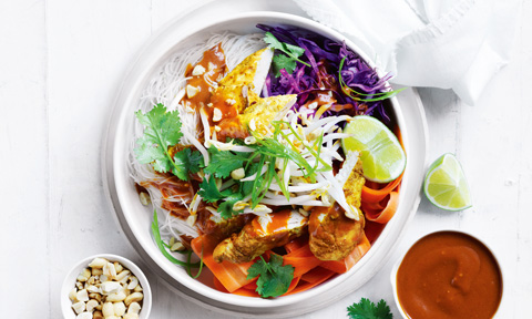 A bowl of satay chicken with noodles, vegetables and a side of nuts
