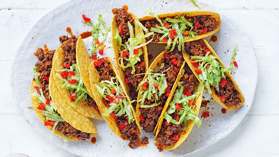 8 beef tacos on a plate topped with shredded lettuce and red capsicum. 