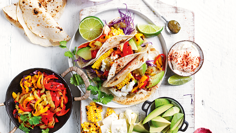 Three tacos served on a plate with corn, avocado, lime wedges and chillies on the side