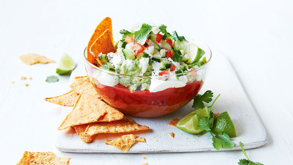 Mexican dip in a bowl topped with corn chips, cucumber, tomato, coriander and lime wedges