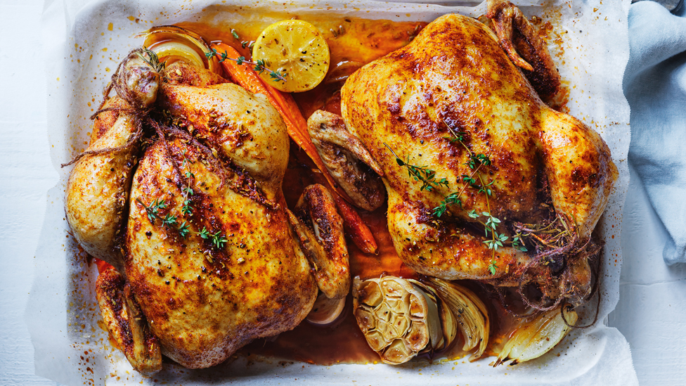 Two roast chickens in a baking tray with lemon, carrot, garlic and thyme