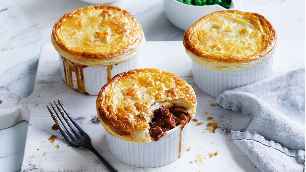 Three beef and guiness pies served in ramekins on a wooden chopping board with a side of peas