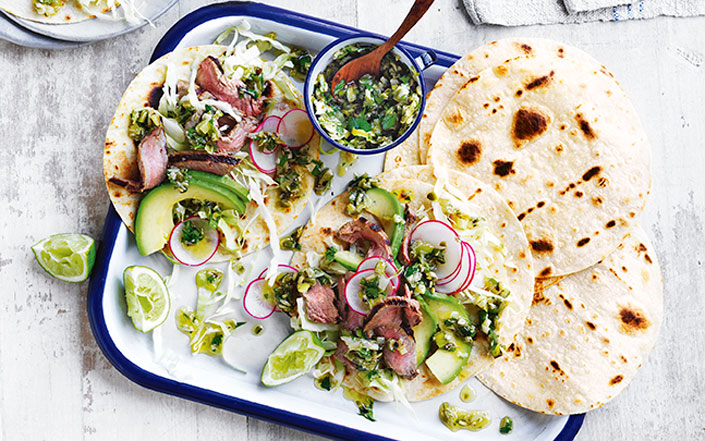 Two steak tacos in a tray garnished with radish, avocado and lime wedges