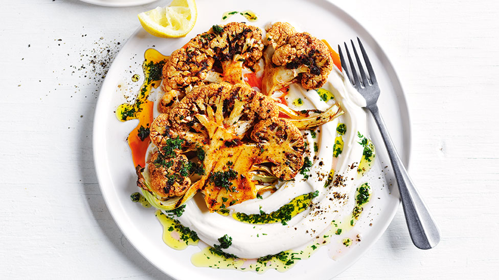 Cauliflower steak served on a plate with yoghurt and chimichurri and lemon wedges