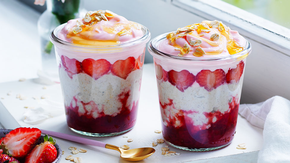 Two glass cups full of strawberry and coconut bircher muesli served alongside spoons and extra strawberries