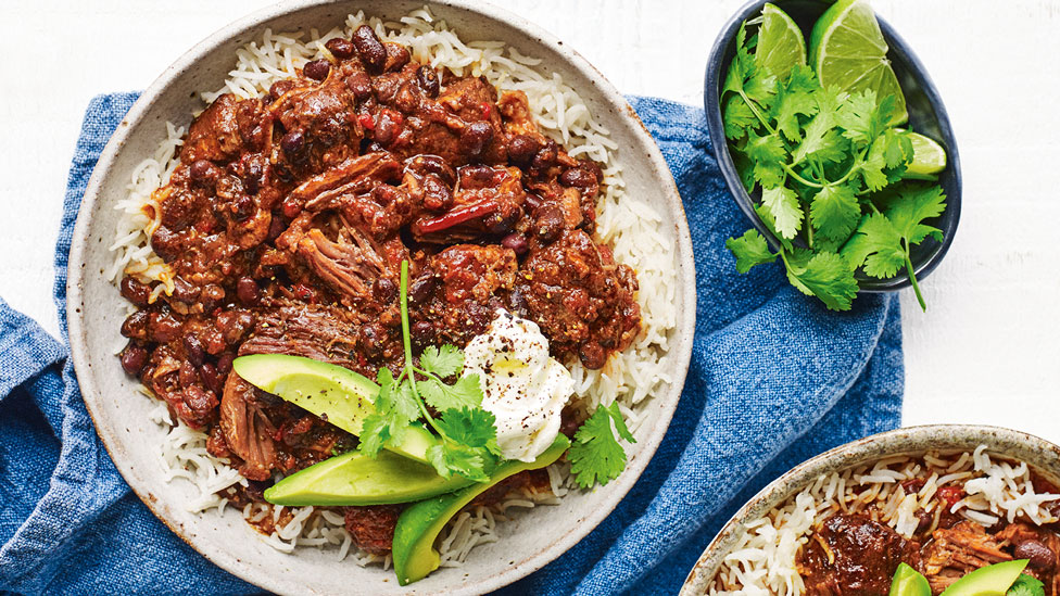 Beef and black bean chilli served on rice with avocado garnish and lime wedges and coriander on the side