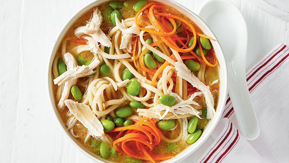 Chicken and udon noodle soup served in a bowl topped with shaved carrot and edamame