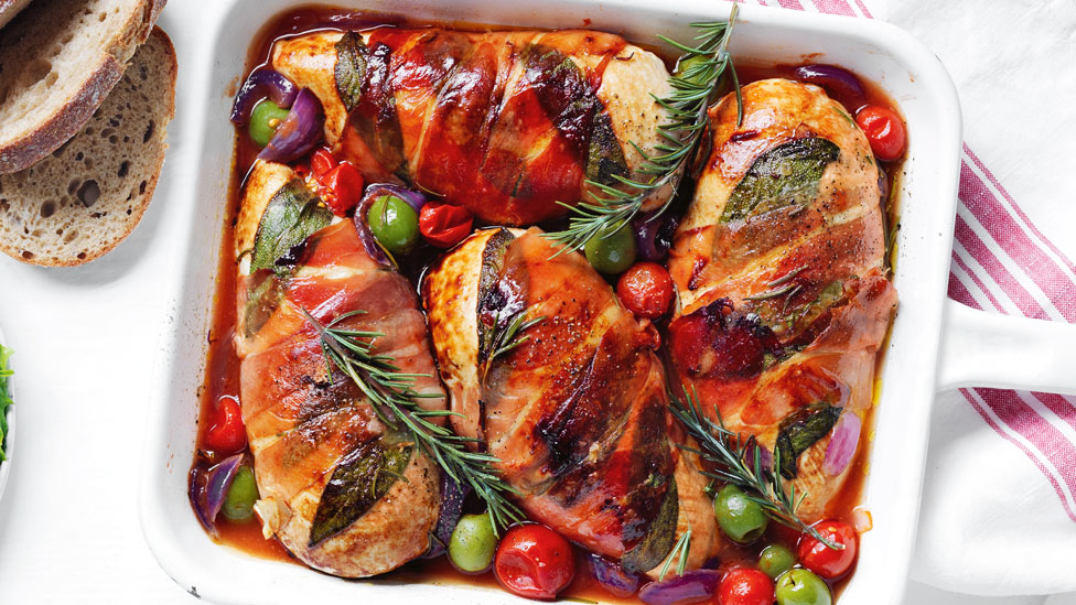 Baking dish with 4 baked chicken pieces wrapped in prosciutto in a tomato sauce, topped with olives, roasted tomatoes and rosemary sprigs