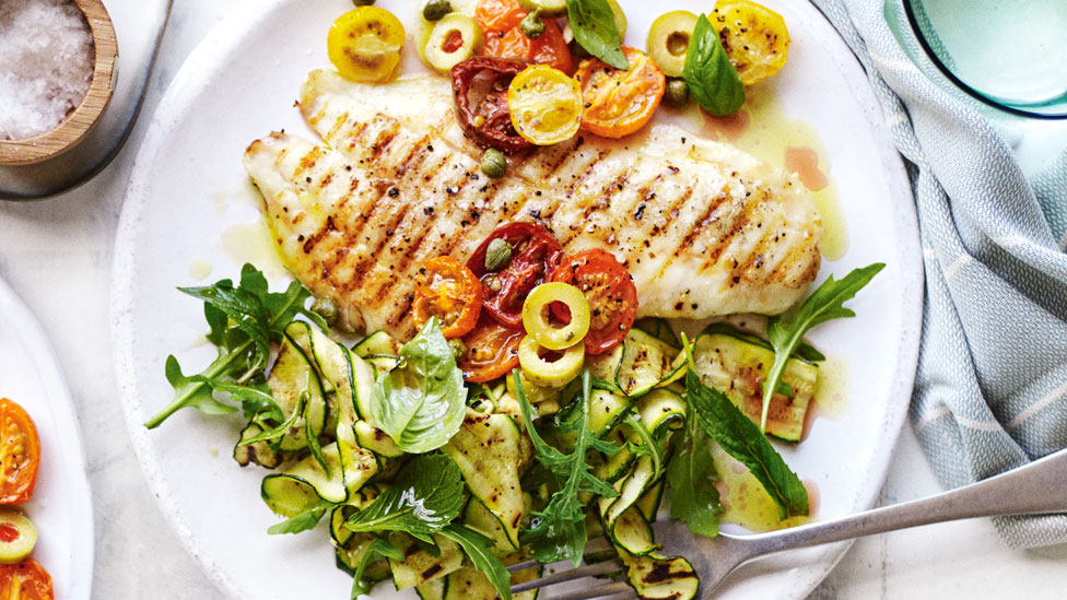 Barbeque Snapper with Roasted Tomato and Caper Relish, served with olived, rocket, basic and mint leaves. Arranged on a table with grey towel and side of coarse salt.