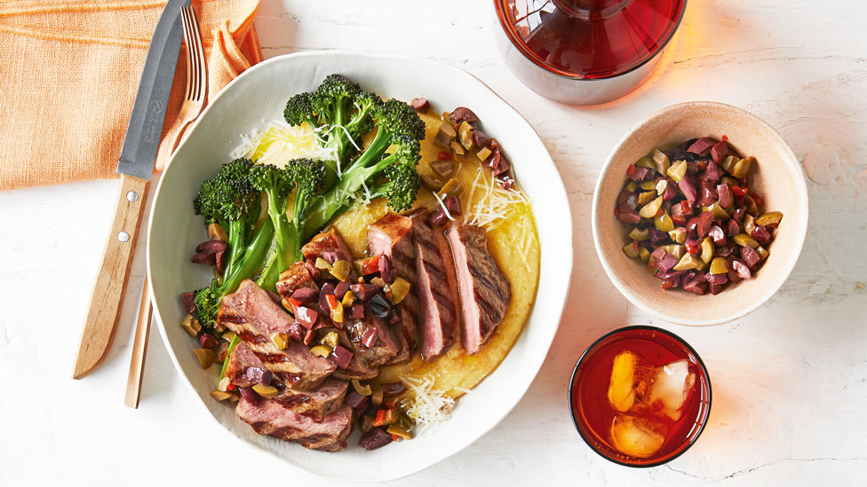 Chargrilled sliced beef with on a bed of polenta with broccoli and grated Parmesan cheese, with a side bowl of chopped olives