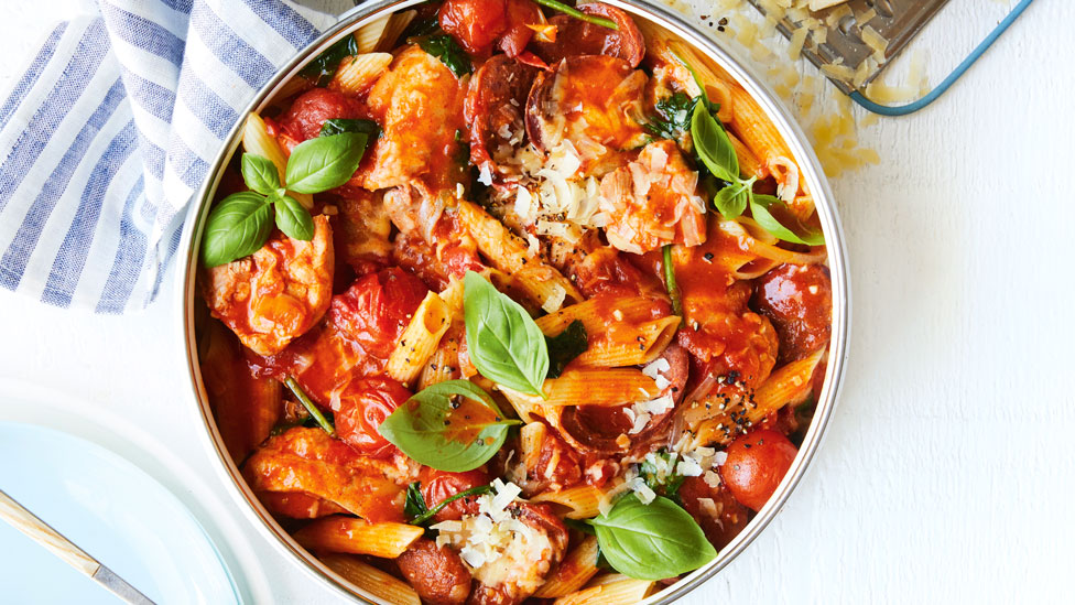 Chicken, Chorizo and Tomato Penne pasta in dish with basil leaves, grated cheese on the side and a striped cloth