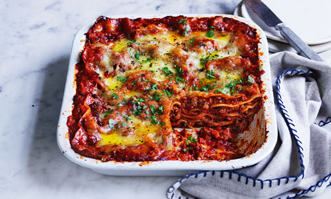 Curtis Stone's Easy Lasagne in white oven dish, topped with parsley. Arranged on marble table with a grey cloth.