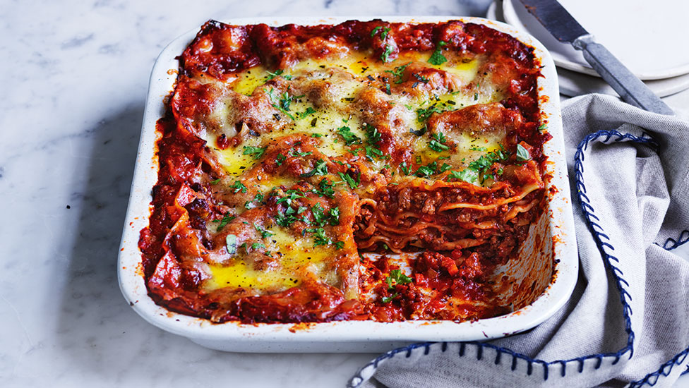 Curtis Stone's Easy Lasagne in white oved dish, topped with parsley. Arranged on marble table with a grey cloth.