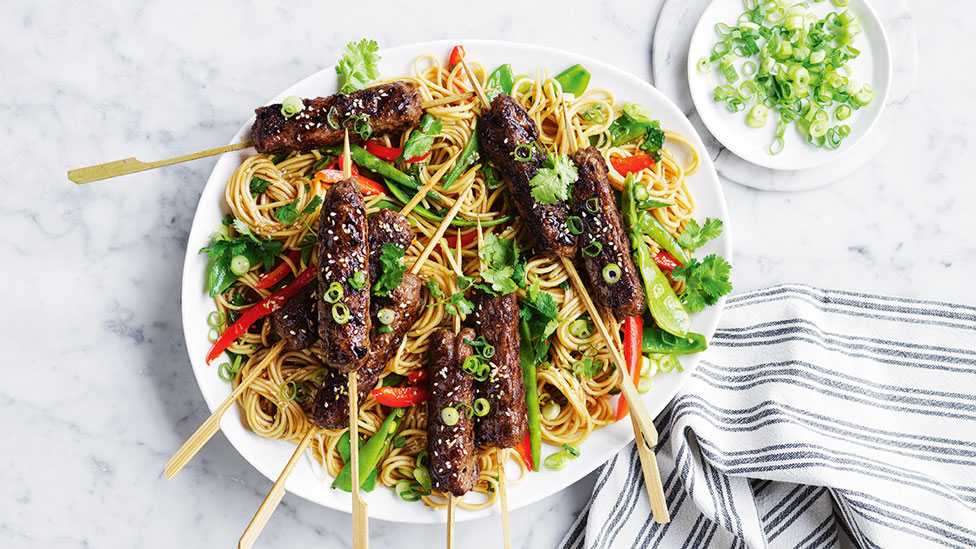  Korean Beef Skewers on Noodles, placed on serving plate on marble table. Spring onion on the dish and on side plate, arranged next to a striped towel.