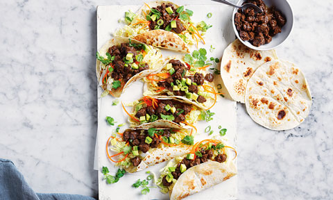 Korean Tacos arranged on baking paper on a marble table. Garnished with avocado, carrot, spring onion and coriander. Additional tacos and mince to the side.