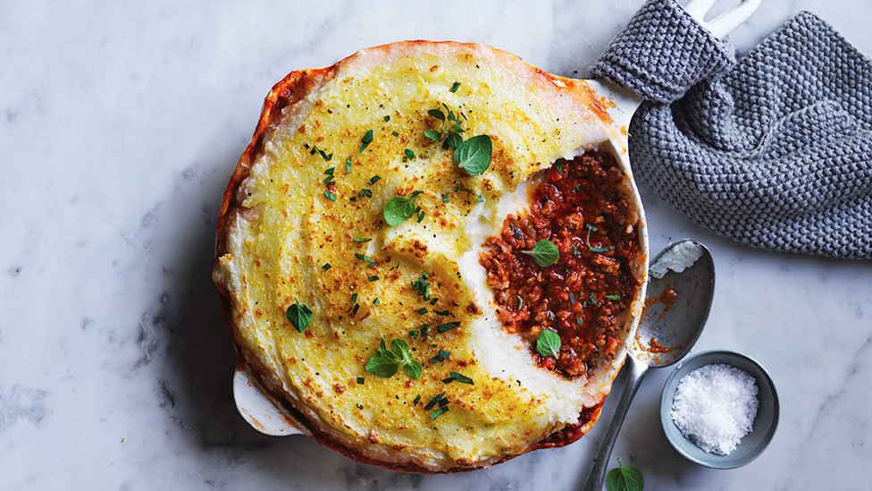 Curtis Stone's Nonna's Cottage Pie garnished with oregano leaves in an oven dish. Salt to the side, arranged on a marble table with a grey towel. 