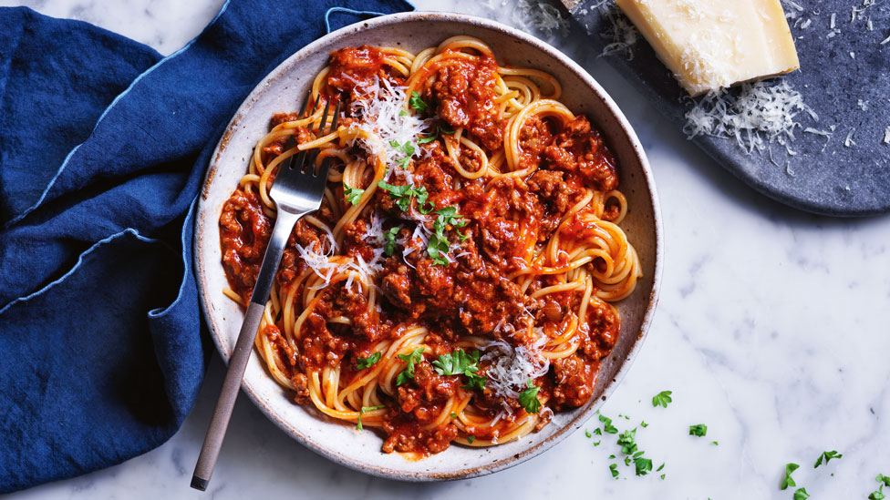 Curtis Stone's Spaghetti Bolognese served in a ceramic bowl, topped with parsley and parmesan. A blue table cloth is on a marble background, a block of parmesan on a navy plate.