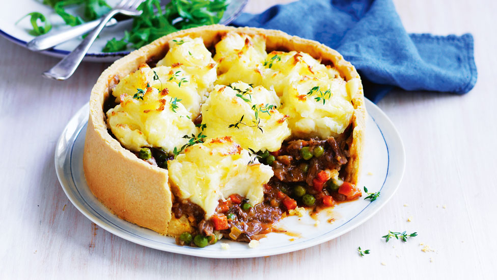 Lamb Shank Shepherd’s Pie on a white plate, topped with thyme sprigs. A plate with rocket is behind, next to a blue cloth.